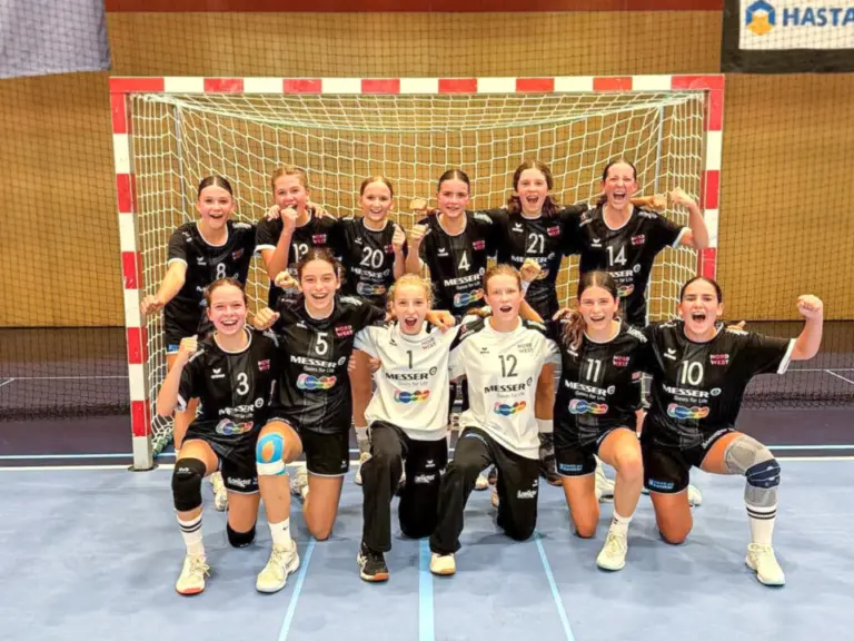 Handballmannschaft mit Mädchen in schwarzen Trikots posiert jubelnd vor einem Tor in einer Sporthalle.
