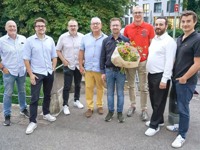Acht Männer stehen nebeneinander im Freien, einer hält einen bunten Blumenstrauss.