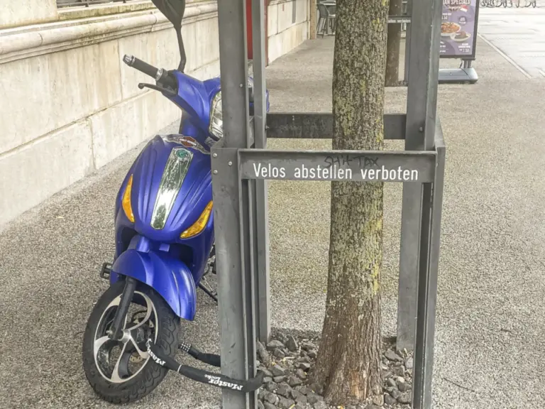 Ein blauer Motorroller steht an einem Baumschutzgitter mit Schild „Velos abstellen verboten“.