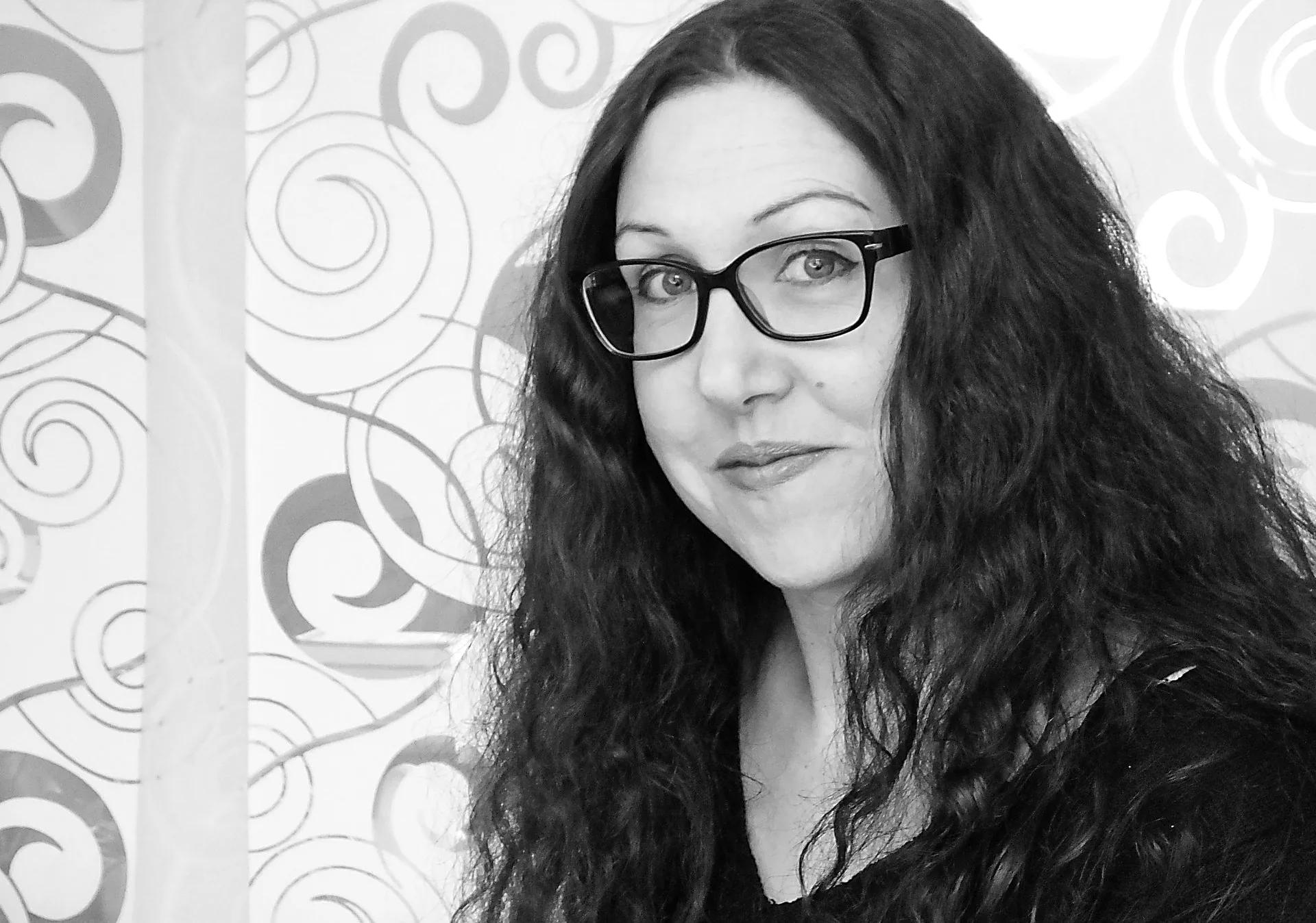 Schwarz-Weiß-Porträt einer lockig-langhaarigen Frau mit Brille vor gemustertem Hintergrund.