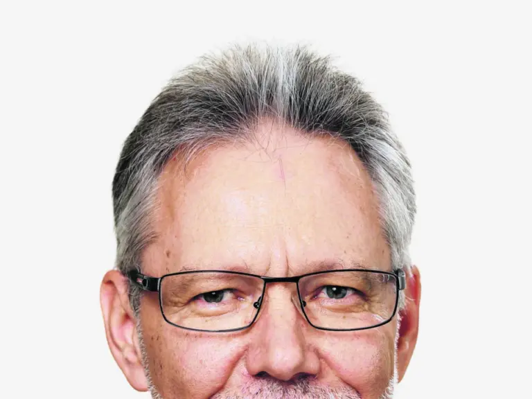 Ein älterer Mann mit grauen Haaren, Brille und Bart lächelt, trägt Anzug mit Krawatte und Anstecknadel.