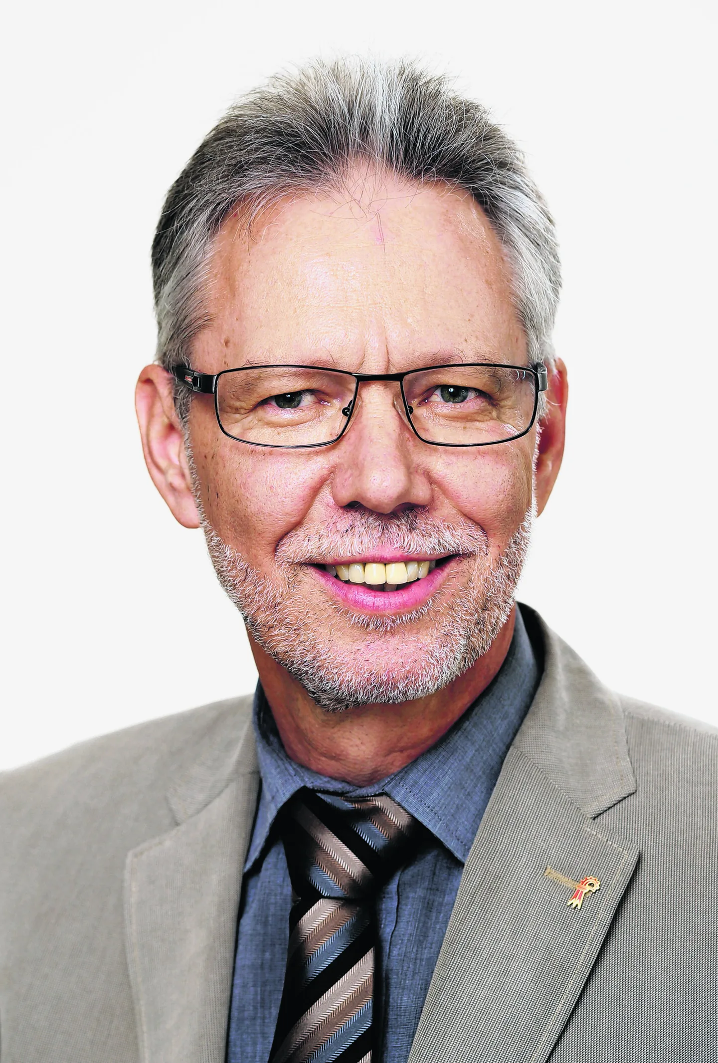Ein älterer Mann mit grauen Haaren, Brille und Bart lächelt, trägt Anzug mit Krawatte und Anstecknadel.