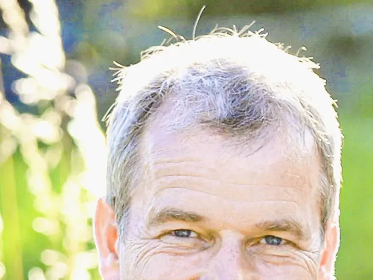 Ein lächelnder Mann mit grauem Haar und hellblauem Hemd steht vor grünem Hintergrund im Freien.