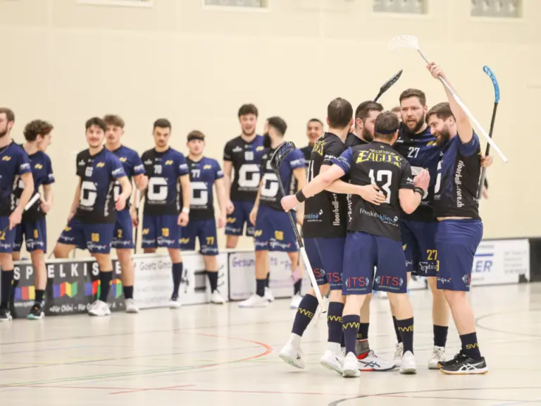Unihockeyspieler in dunklen Trikots umarmen sich jubelnd in einer Sporthalle, im Hintergrund weitere Spieler und ein Schiedsrichter.