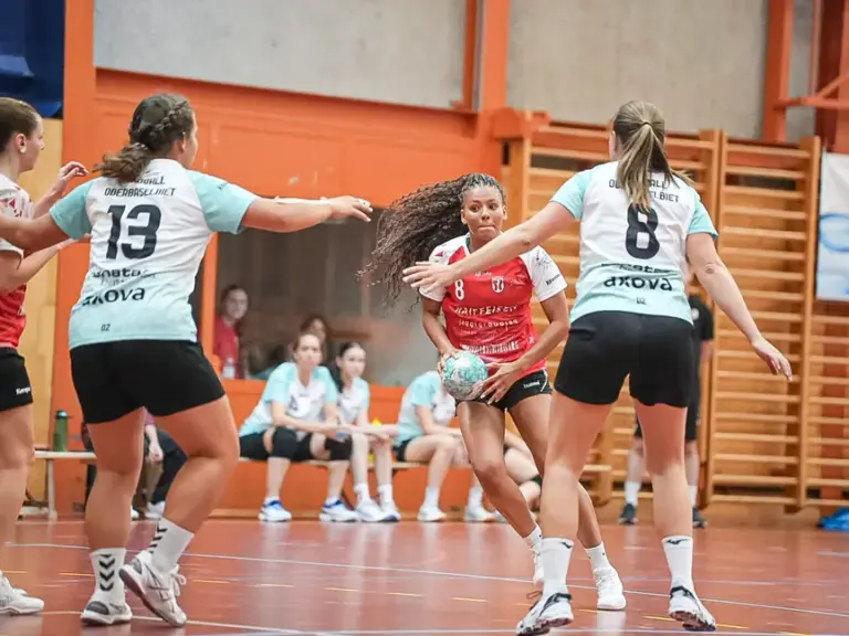Handballspielerinnen im Zweikampf um den Ball in einer Sporthalle.