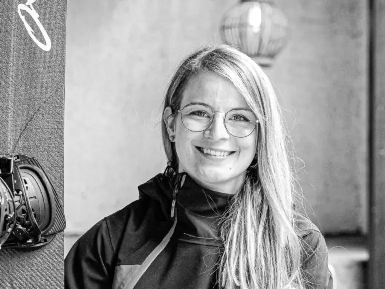 Lächelnde Frau mit blondem Haar sitzt neben einem Snowboard auf einer Holzbank im Freien.