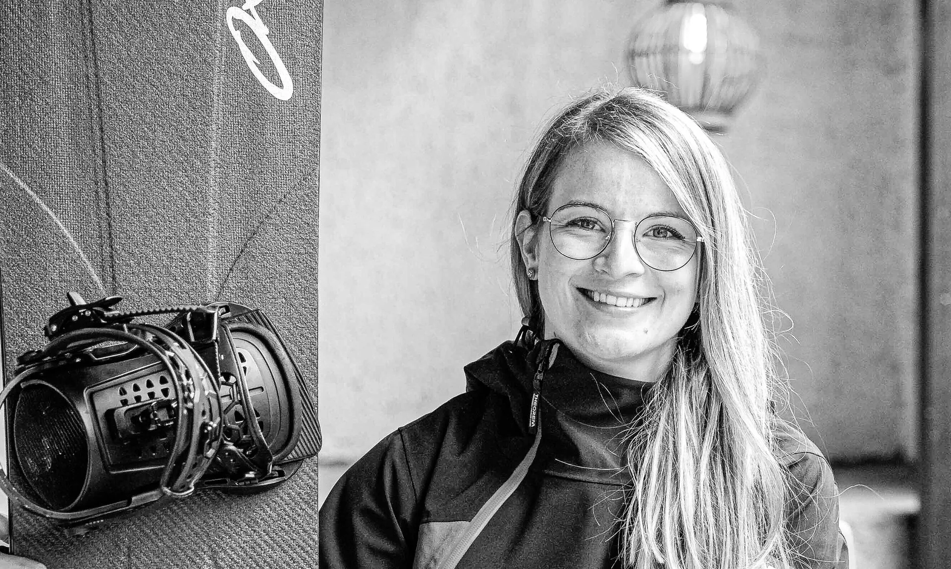 Lächelnde Frau mit blondem Haar sitzt neben einem Snowboard auf einer Holzbank im Freien.