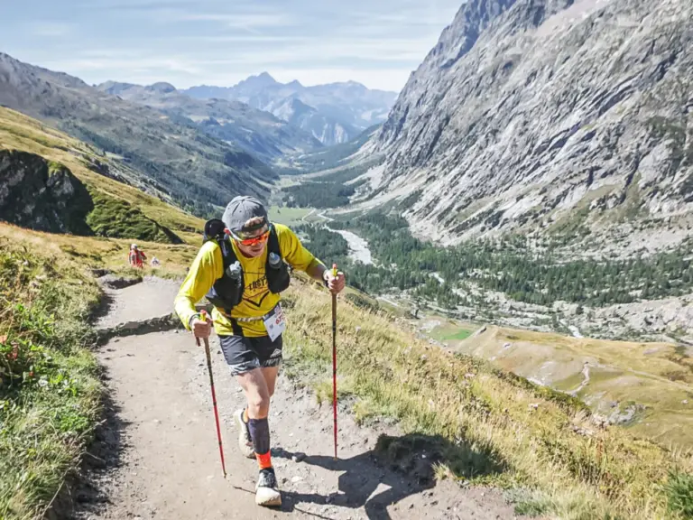 Ein Läufer mit Trekkingstöcken und Rucksack steigt einen steilen Bergpfad hinauf, im Hintergrund ein weites Alpental.