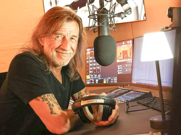 Ein lächelnder Mann mit langen Haaren sitzt vor einem Mikrofon in einem Radiostudio, Kopfhörer in der Hand.