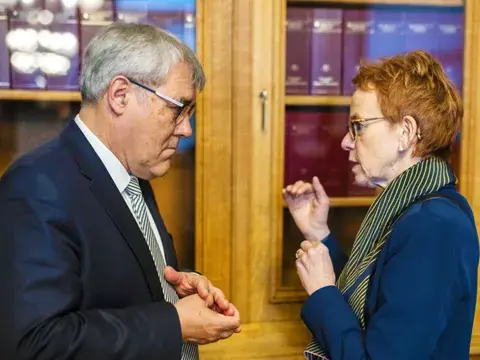 Ein Mann und eine Frau stehen sich gegenüber und sprechen miteinander, im Hintergrund Bücherregale.