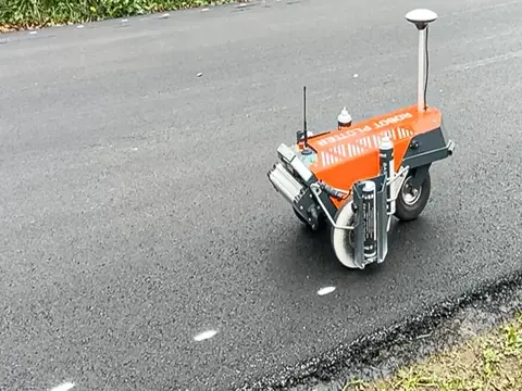 Ein oranger Roboter mit Antennen und Rädern steht auf einer frisch asphaltierten Strasse und setzt weisse Markierungspunkte.