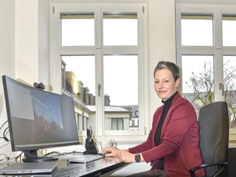 Eine Frau in roter Jacke sitzt an einem Schreibtisch mit Computer und blickt lächelnd zur Seite.