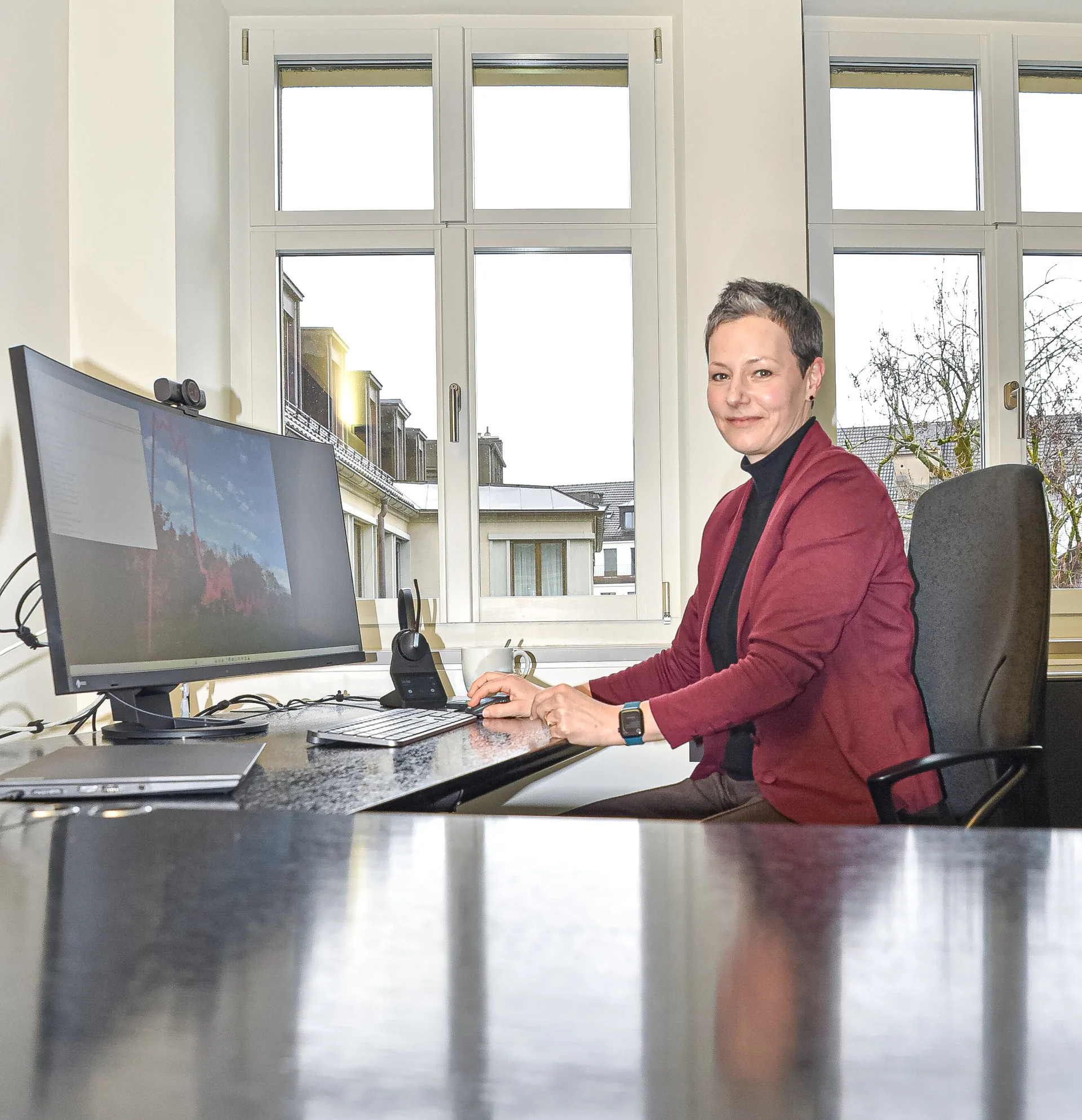 Eine Frau in roter Jacke sitzt an einem Schreibtisch mit Computer und blickt lächelnd zur Seite.