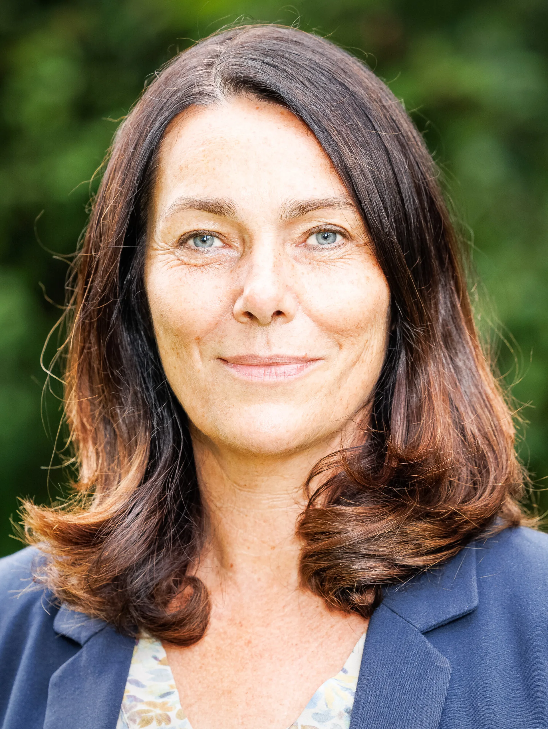 Eine Frau mittleren Alters mit schulterlangen braunen Haaren lächelt leicht vor grünem Hintergrund.