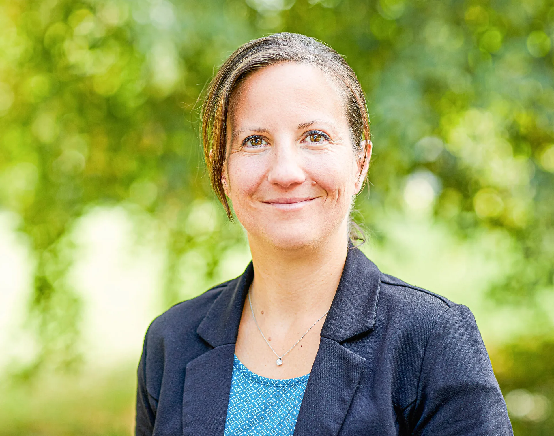 Eine Frau mittleren Alters mit zurückgebundenem Haar lächelt vor grünem Blätterhintergrund, sie trägt einen dunklen Blazer.