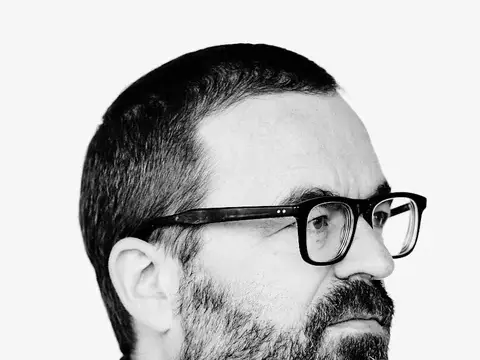 Schwarz-weiß-Porträt eines Mannes mit Bart und Brille im Profil, dunkle Jacke, heller Hintergrund.