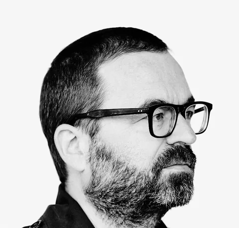 Schwarz-weiß-Porträt eines Mannes mit Bart und Brille im Profil, dunkle Jacke, heller Hintergrund.
