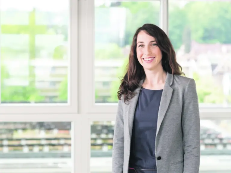 Eine lächelnde Frau mit langen dunklen Haaren im grauen Blazer steht vor grossen Fenstern.