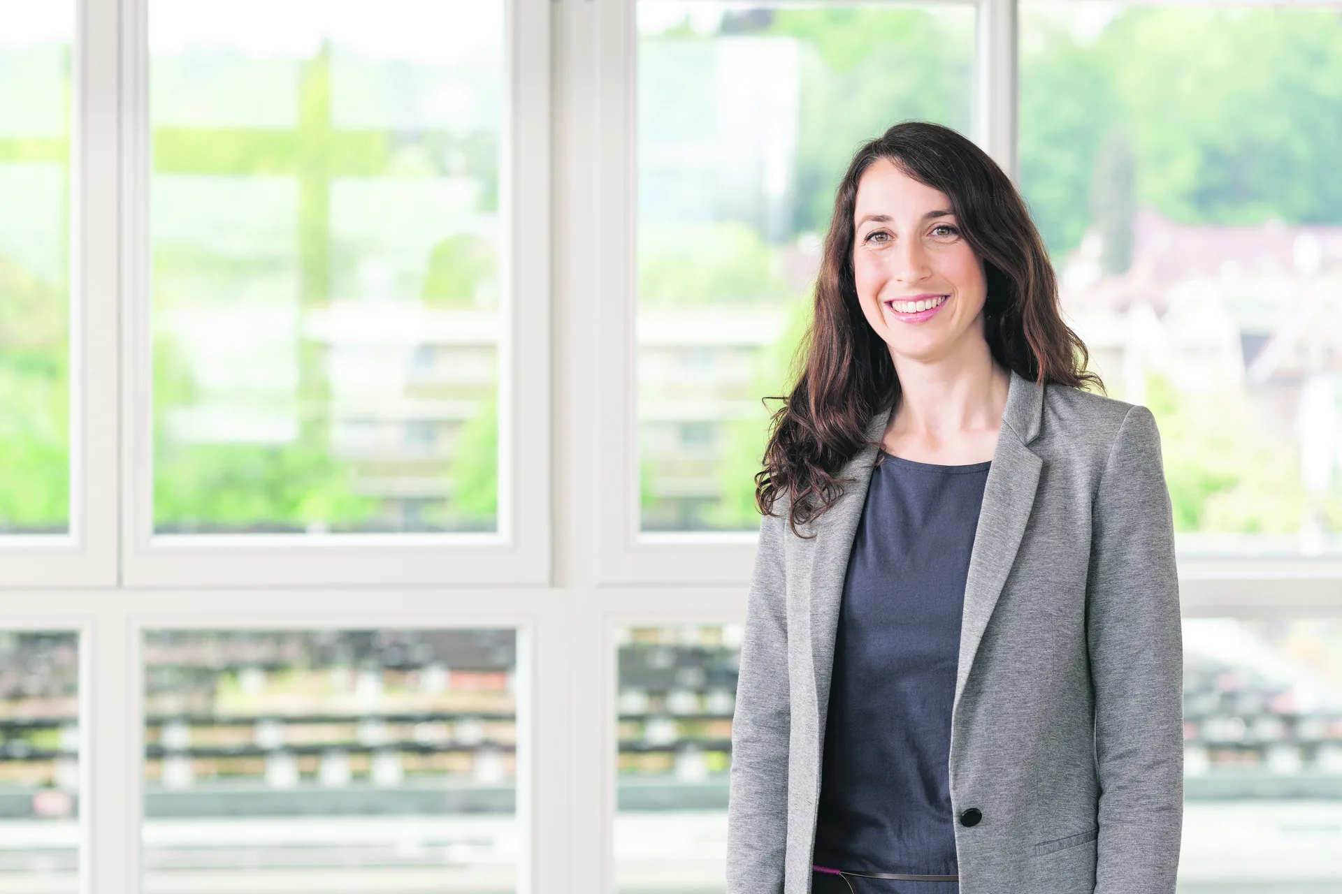Eine lächelnde Frau mit langen dunklen Haaren im grauen Blazer steht vor grossen Fenstern.