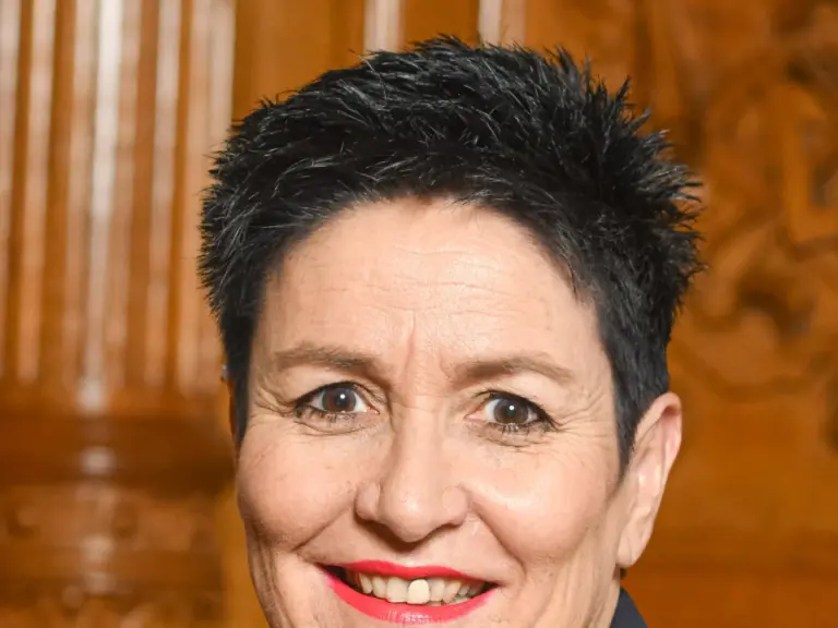 Lächelnde Frau mit kurzen dunklen Haaren in dunklem Blazer vor einer geschnitzten Holzwand.