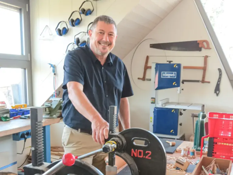 Ein lächelnder Mann steht in einer Werkstatt an einer Presse mit der Aufschrift „No.2“, im Hintergrund Werkzeuge und Holzspäne.