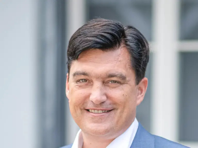 Ein lächelnder Mann im blauen Sakko steht vor einem hellen Gebäude mit Sprossenfenstern.