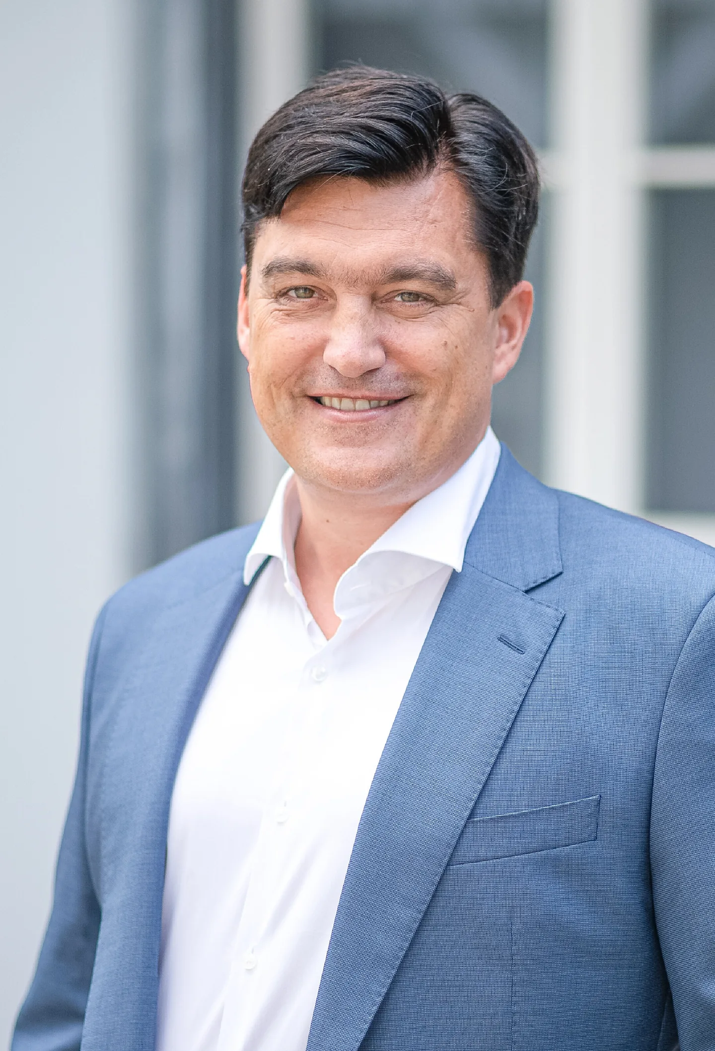 Ein lächelnder Mann im blauen Sakko steht vor einem hellen Gebäude mit Sprossenfenstern.