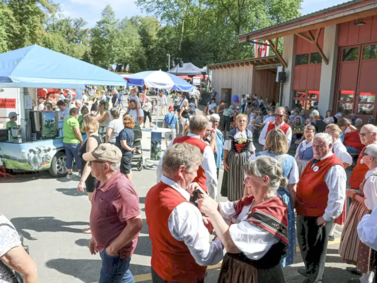 Menschen in Trachten und Alltagskleidung unterhalten sich auf einem sonnigen Marktplatz mit Zelten.