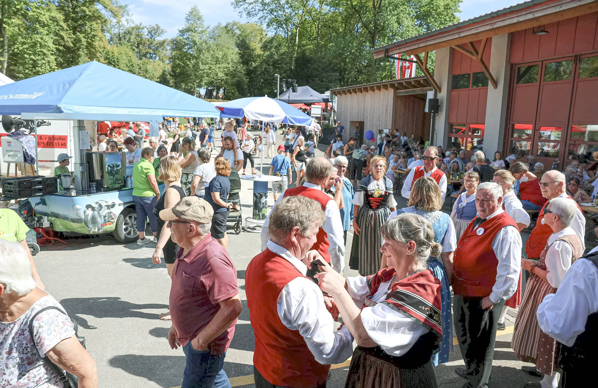 Menschen in Trachten und Alltagskleidung unterhalten sich auf einem sonnigen Marktplatz mit Zelten.