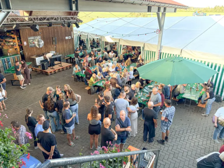 Menschenmenge steht und sitzt an Bierbänken unter einem Zeltdach und einem grünen Sonnenschirm im Freien.