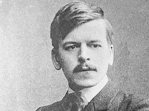 Historisches Porträtfoto eines jungen Mannes mit Schnurrbart, Anzug und verschränkten Armen.