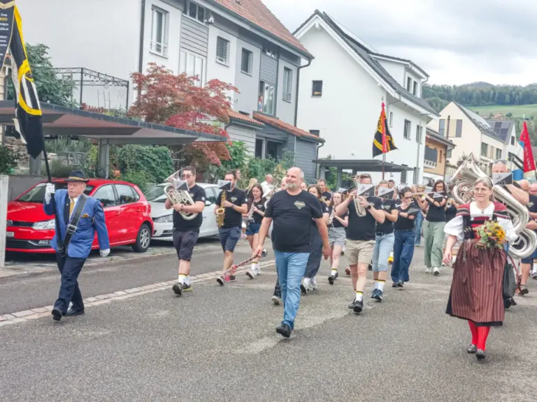 Musikkapelle mit Blasinstrumenten und Fahnenträgern marschiert durch eine Wohnstrasse, eine Frau in Tracht voran.
