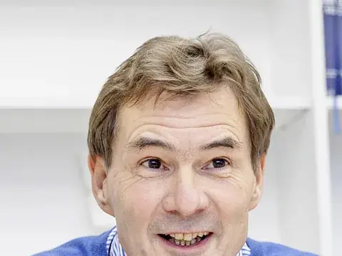Ein Mann in blauem Pullover sitzt an einem Schreibtisch, gestikuliert lächelnd mit einem Stift in der Hand.