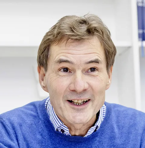 Ein Mann in blauem Pullover sitzt an einem Schreibtisch, gestikuliert lächelnd mit einem Stift in der Hand.