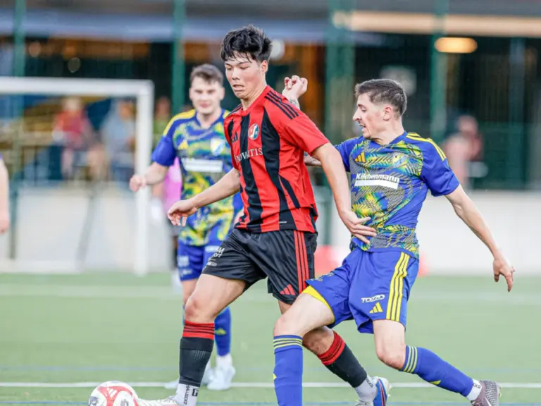 Zwei Fussballspieler kämpfen auf dem Rasen um den Ball, im Hintergrund ein weiterer Spieler.