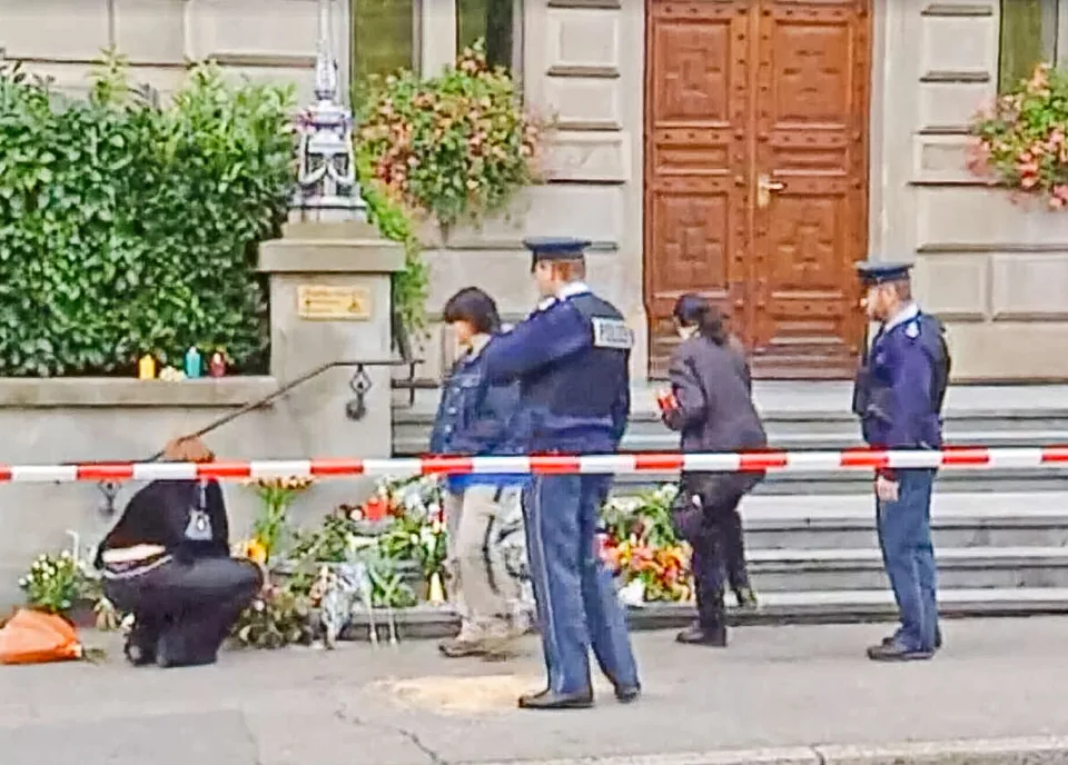 Polizisten und Passanten stehen vor einem Gebäudeeingang bei Blumen und Kerzen hinter Absperrband.