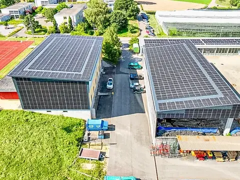 Luftaufnahme zweier Industriehallen mit grossen Photovoltaikanlagen auf den Dächern, umgeben von Feldern.