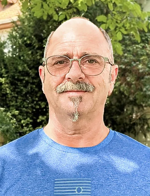 Ein älterer Mann mit Brille, Schnauzbart und Glatze lächelt in einem blauen T-Shirt vor grünen Hecken.
