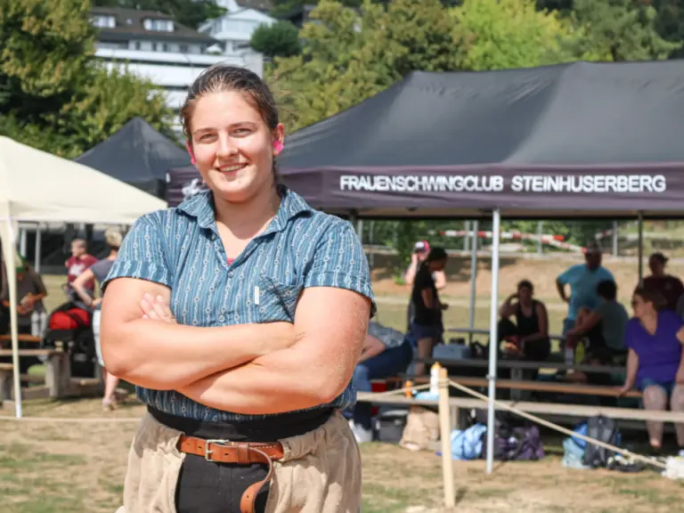 Eine junge Frau mit verschränkten Armen steht lächelnd im Freien vor einem Festzelt.