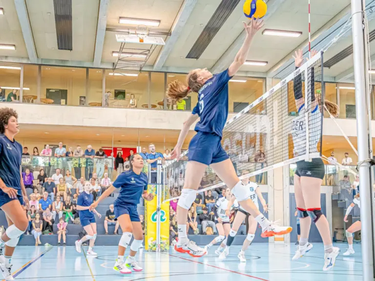 Volleyballspielerin springt zum Angriffsschlag am Netz, umgeben von Mitspielerinnen und Zuschauern in der Halle.