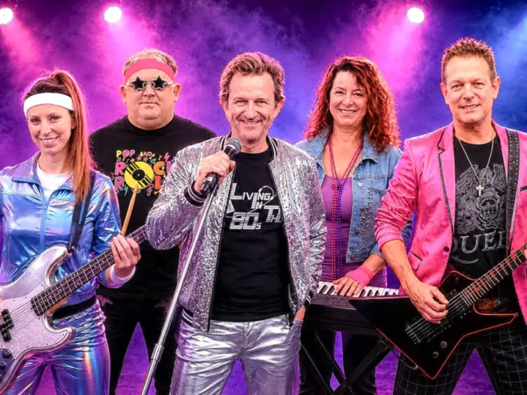 Fünf Musiker in bunten 80er-Jahre-Outfits stehen mit Instrumenten und Mikrofon vor buntem Bühnennebel.