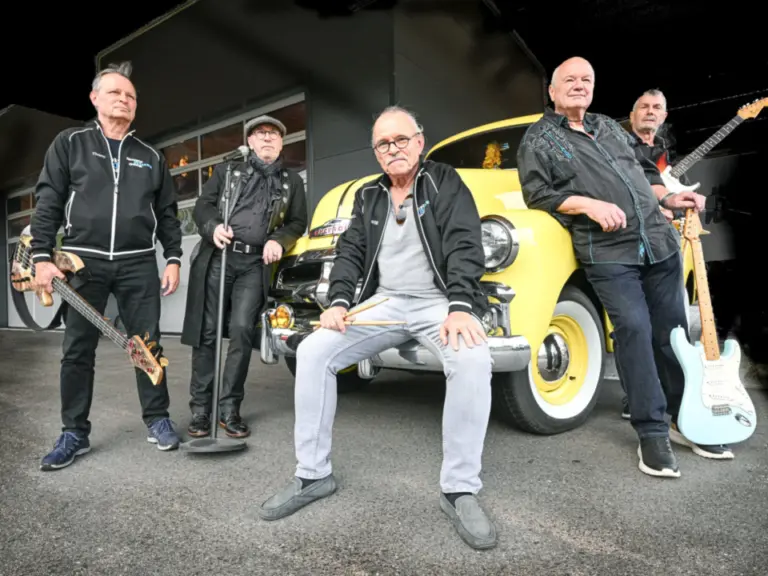Fünf ältere Männer mit Gitarren und Mikrofon posieren vor einem gelben Oldtimer-Auto.