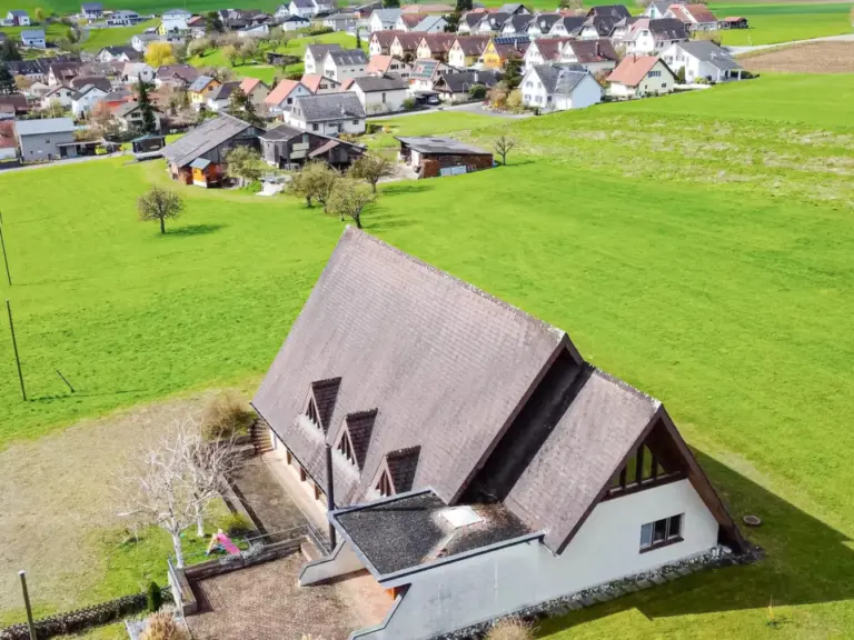 Luftaufnahme eines Gebäudes mit spitzem Satteldach, umgeben von grünen Wiesen und einem Dorf im Hintergrund.