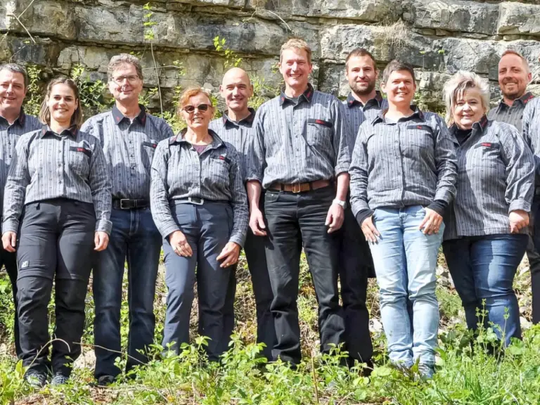Eine Gruppe von etwa fünfzehn Personen in einheitlichen grauen Hemden steht nebeneinander vor einer Felswand.