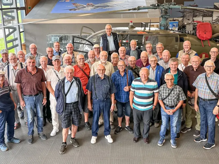 Grosse Gruppe älterer Männer posiert in einem Flugzeughangar vor einem Helikopter.