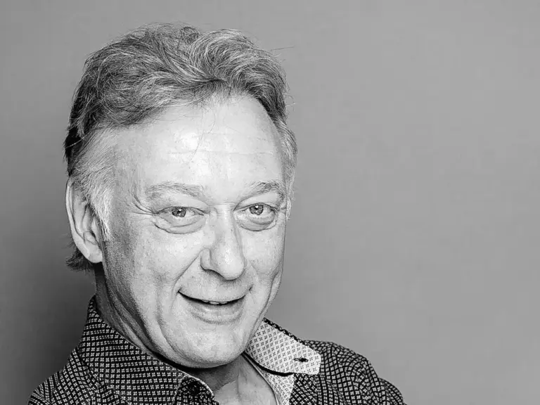 Ein älterer lächelnder Mann mit verschränkten Armen in gemustertem Hemd, Schwarz-Weiß-Porträt.