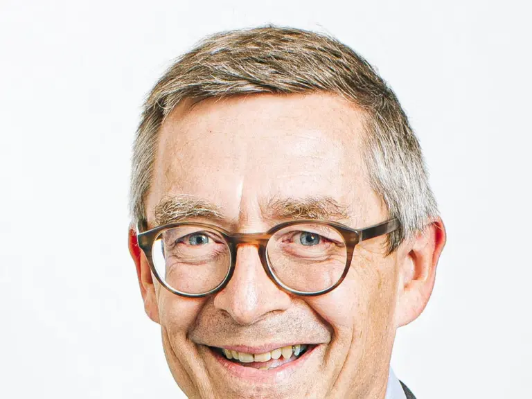 Ein lächelnder älterer Mann mit Brille, grauem Haar und dunklem Sakko vor hellem Hintergrund.