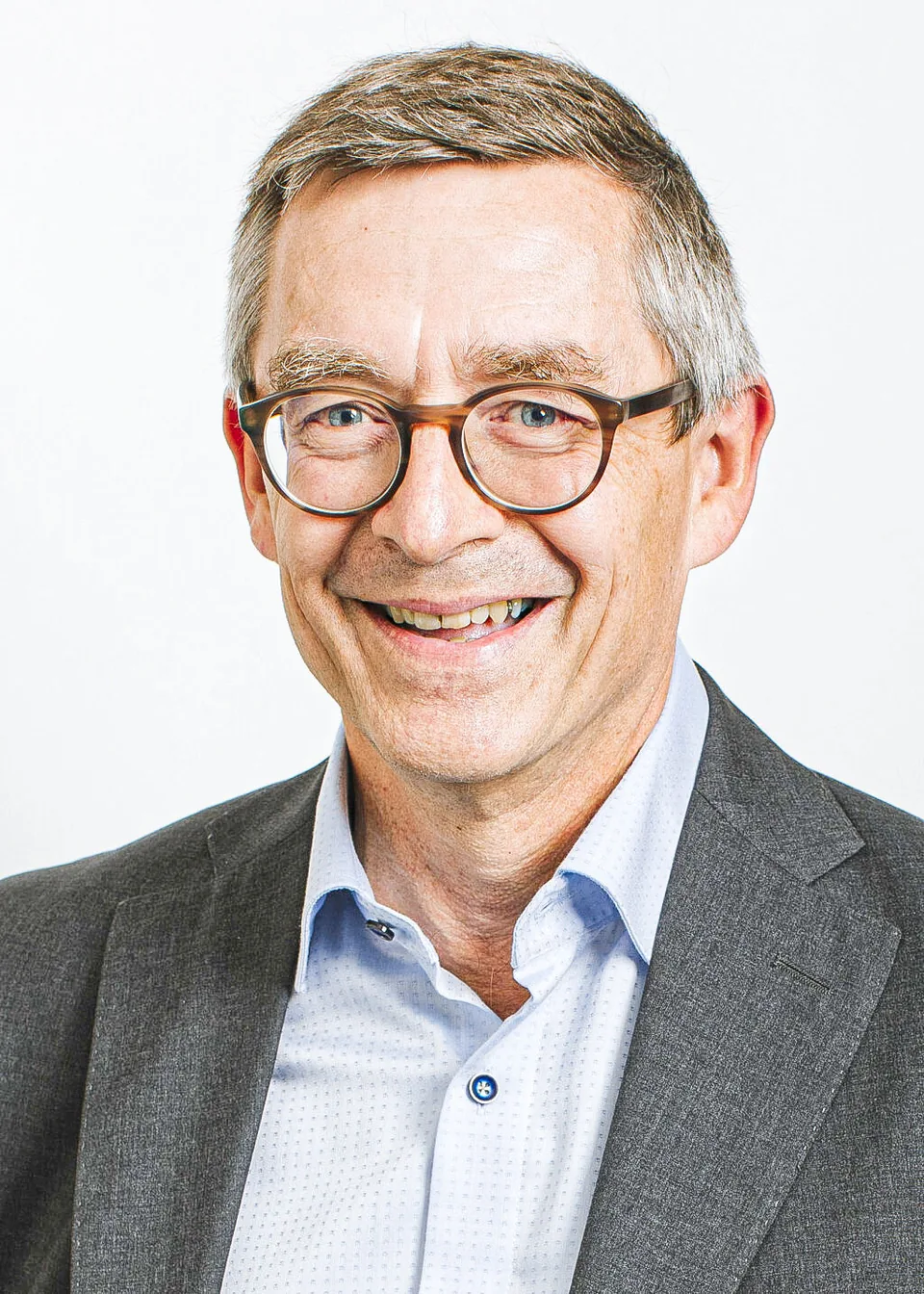 Ein lächelnder älterer Mann mit Brille, grauem Haar und dunklem Sakko vor hellem Hintergrund.