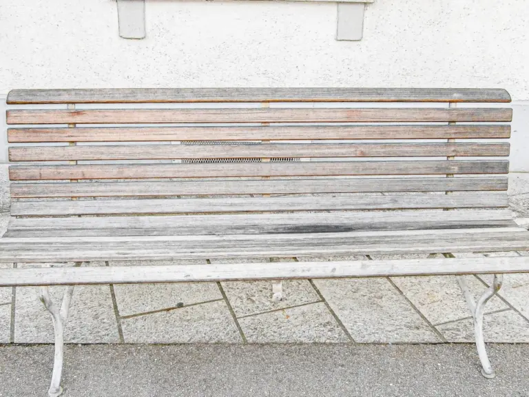Eine hölzerne Sitzbank mit weissem Metallgestell steht auf Steinplatten vor einem Gebäude.