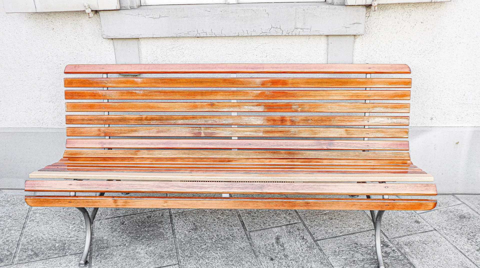 Eine leere Holzbank mit geschwungenen Metallbeinen steht auf gepflasterten Steinplatten vor einer Hausfassade.
